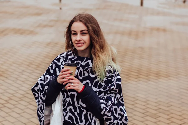 レインコートを着たスタイリッシュな女性が市街地を歩き テイクアウトコーヒーを手に持つ — ストック写真