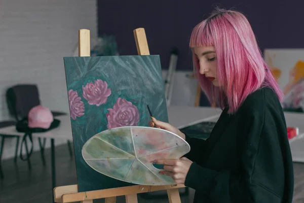 Gorgeous female artist with pink hair working on art projects with palette in her hands in the art studio. Young woman draws a picture