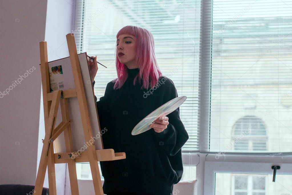 Gorgeous female artist with pink hair working on art projects with palette in her hands in the art studio. Young woman draws a picture
