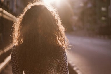 Mutlu kıvırcık genç kadın açık havada gülümseyen ve gün batımında şehir caddesinde yürüyüş 