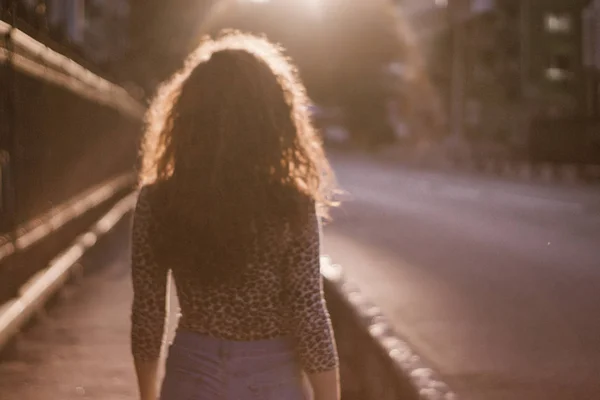 Mutlu kıvırcık genç kadın açık havada gülümseyen ve gün batımında şehir caddesinde yürüyüş 