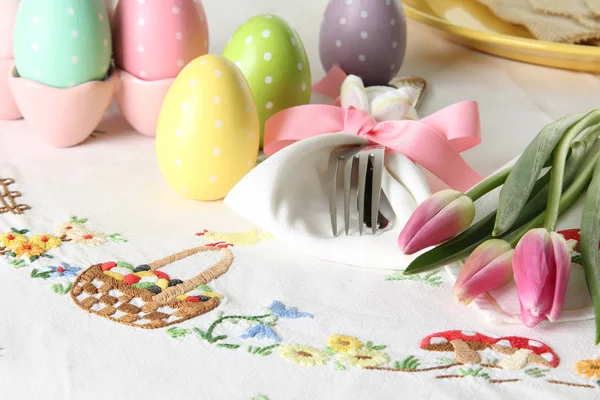 この伝統的な祝日のブランチの場所の設定に塗られた卵 刺繍のイースター バスケット ナプキンと春ピンクのチューリップに包まれた銀のカトラリーが含まれています ストック画像