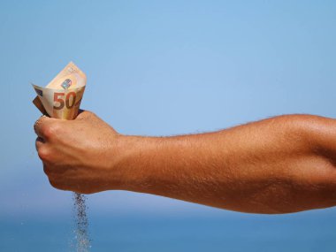 male arm holds fifty euro banknote and lets sand trickle through on blue sea background, concept of holiday cost, expensive holiday, money turns to sand