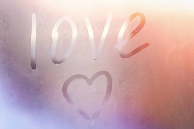 Terli bardağa kazınmış aşk ve kalp. Renkli arka planda sisli cam bir pencereye çizilmiş aşk kalbi. Sisli camda aşkın ve mutluluğun sembolü.