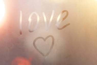 Terli bardağa kazınmış aşk ve kalp. Renkli arka planda sisli cam bir pencereye çizilmiş aşk kalbi. Sisli camda aşkın ve mutluluğun sembolü.