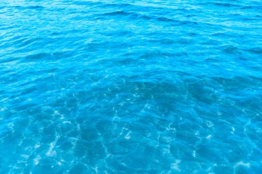 Sahilin arka planında mavi şeffaf deniz suyu. Güneşli bir günde hafif dalgalar ve parıltılarla berrak su yüzeyi