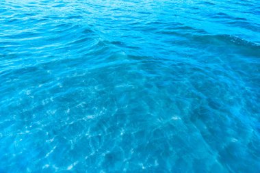 Sahilin arka planında mavi şeffaf deniz suyu. Güneşli bir günde hafif dalgalar ve parıltılarla berrak su yüzeyi