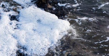 Sahilde beyaz köpük birikintileri var. Su yüzeyi dokusu arka plan, nehir köpüğü, kümülüs köpüğü, hava köpüğü, bej deniz gölü köpüğü, su üzerinde köpüklü kabarcıklar.