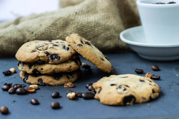 A cookie with chocolate drops lies on a stone board with coffee and peanuts scattered around the grains. There is a burlap next to it and there is a white cup with coffee beans