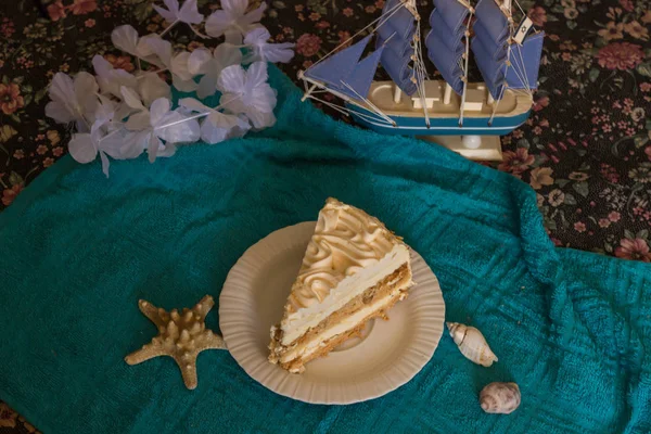 A piece of an airy Kiev cake on a white plate with a marine decor in the form of a blue ship and seashells