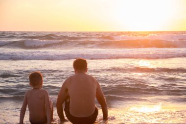 Babası denizde kum üzerine oturan oğlu günbatımı gösterir