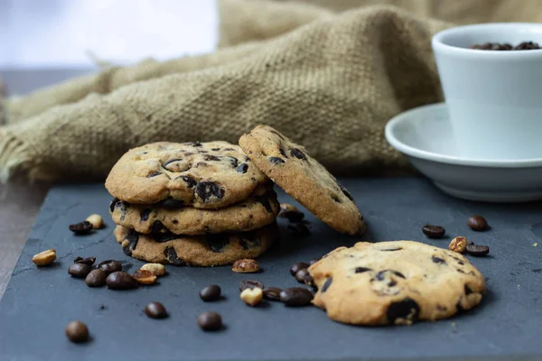 A cookie with chocolate drops lies on a stone board with coffee and peanuts scattered around the grains. There is a burlap next to it and there is a white cup with coffee beans