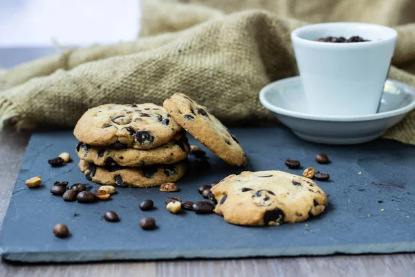 A cookie with chocolate drops lies on a stone board with coffee and peanuts scattered around the grains. There is a burlap next to it and there is a white cup with coffee beans