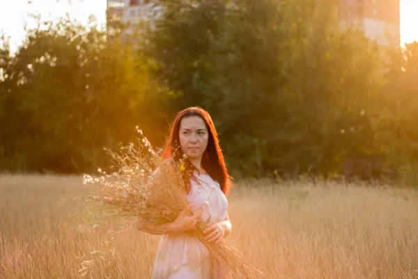 Bella ragazza in un lungo abito leggero sta divertendosi, ballando e raccogliendo fiori selvatici in un tramonto arancione — Foto Stock