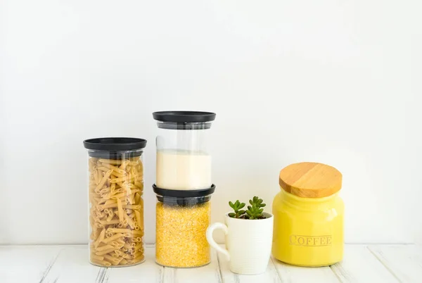 Pasta, coffee and cereals in glass jars and a small plant in a cup on white background. Organized home kitchen and healthy eating concept. Minimalist bright composition with place for text