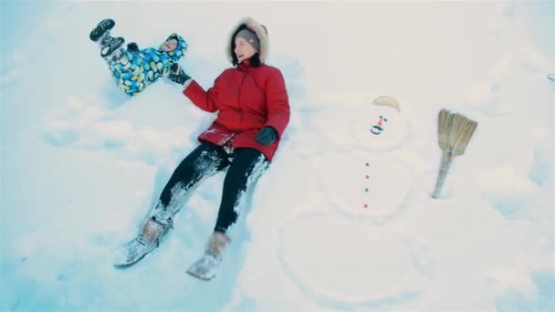 Mãe e filho brincando deitados na neve ao lado de um boneco de neve — Vídeo de Stock