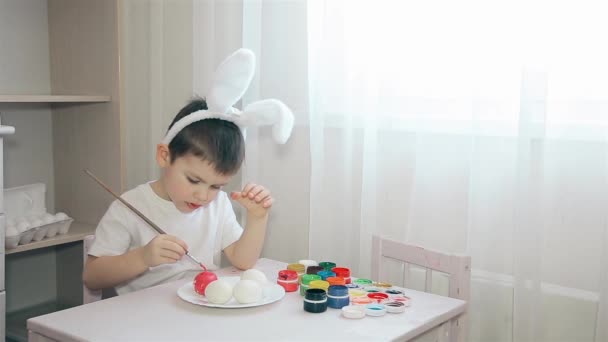 Un bambino con le orecchie di coniglio sulla testa dipinge un uovo di Pasqua in rosa — Video Stock