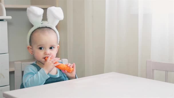 Een klein meisje in een witte Bunny kostuum zit aan de tafel en nibbles een wortel — Stockvideo