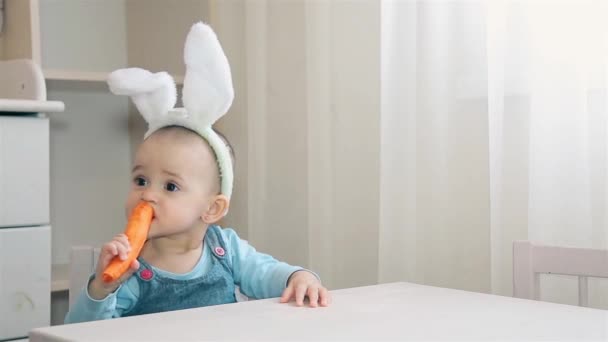 Ребенок дошкольного возраста с заячьими ушами на голове сидит в детской и жует оранжевую морковку — стоковое видео