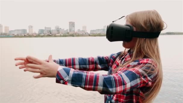 Blond dziewczyna z okularami wirtualna rzeczywistość i koszula w kratę stoi na tle dużego miasta w pobliżu morza — Wideo stockowe