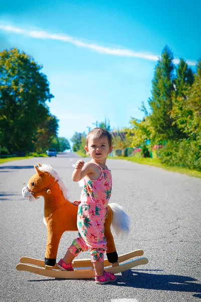A little girl stands alone near a toy horse outdoors on an asphalt road