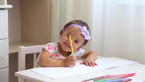 Meisje in bloemenjurk leert tekenen op een stuk papier met gekleurde potloden aan een witte tafel — Stockvideo