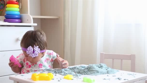 Маленька дівчинка у квітковій сукні за білим столом грає з конічним піском — стокове відео