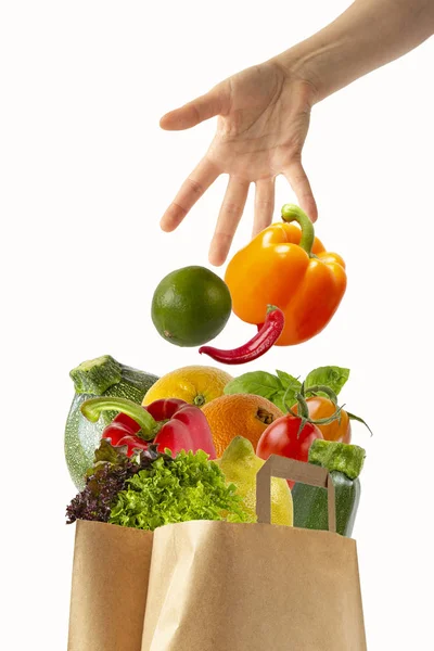 Kvinnlig hand kastar grönsaker i en papperskasse med mat. Isolerade objekt på vit bakgrund. Stockbild