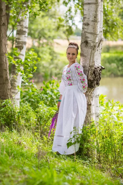 En el campo. Mujer rural en el fondo de la naturaleza en vestidos bordados.Edificios de madera en el pueblo. Campos prados  . — Foto de Stock
