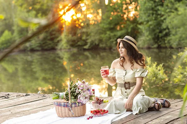 Ung vacker, mörkhårig flicka i en stråhatt vid solnedgången vid sjön på en picknick på fältet. Sommaren. Friluftsliv frukt och dryck i en korg och en bukett — Stockfoto