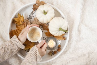 Yakın - bir fincan kahve tutan eller kadar, yanan bir mum, ahşap bir pansuman üzerinde. Battaniyenin üzerindeki postada kahve. Yeşillik ve kabak mevsimlik dekorasyon . Sonbahar