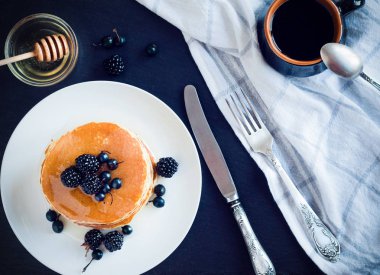 Blackberry ve siyah frenk üzümü, kahve ve koyu arka plan üzerinde bal ile fincan tabağı ile krep yığını üstten görünüm.