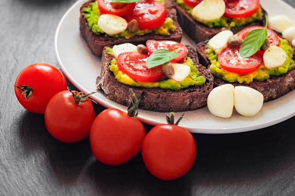 Kiraz domates, mozzarella peyniri, avokado, fesleğen ve zeytinyağı sandviç tost. Karanlık bir taş yemek yan görünüm.