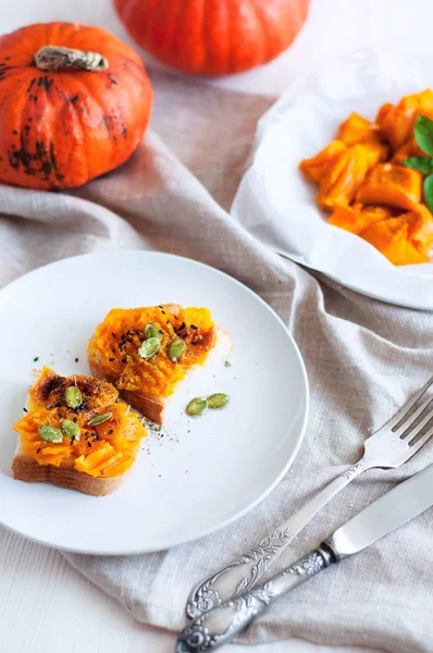 Toast with mashed pumpkin with green, pepper and seeds. Sliced roasted pumpkin on white parchment sprinkled with spices. Angle view still life on wooden and fabric background. — Stock Photo, Image