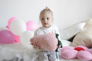 Bir kız beyaz ve pembe balonlarla ve çilek yastıkla yatakta oynuyor.