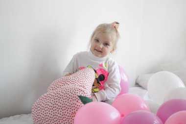 Bir kız beyaz ve pembe balonlarla ve çilek yastıkla yatakta oynuyor.