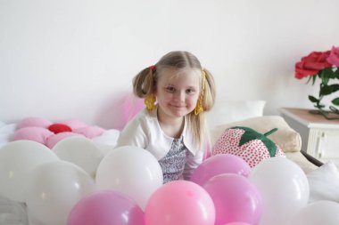 Bir kız beyaz ve pembe balonlarla ve çilek yastıkla yatakta oynuyor.