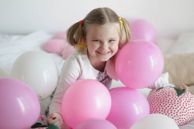 Bir kız beyaz ve pembe balonlarla ve çilek yastıkla yatakta oynuyor.