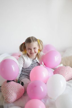 Bir kız beyaz ve pembe balonlarla ve çilek yastıkla yatakta oynuyor.