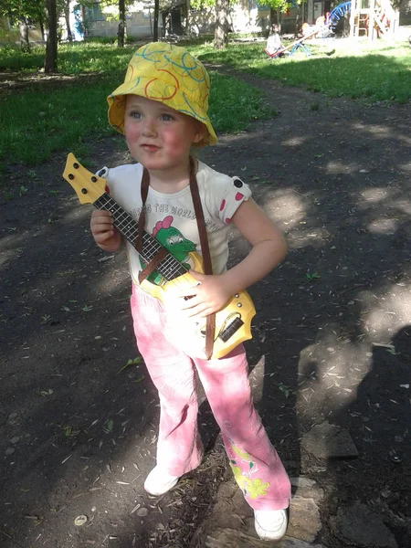 一个穿粉红色裤子的女孩 一件白色的T恤和一顶黄色的帽子在儿童黄色的吉他上演奏 — 图库照片