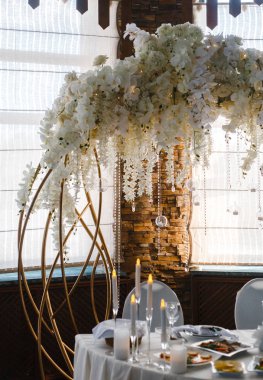 Düğün masasının arkasında beyaz çiçeklerle süslenmiş dekoratif kemer.