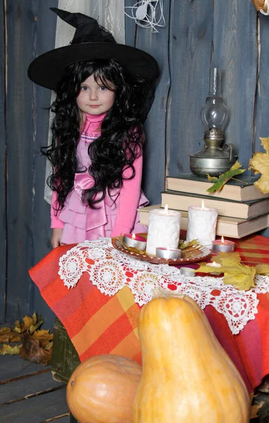 a girl in a witch\'s cap and wig stands near a table with books, candles and a kerosene lamp