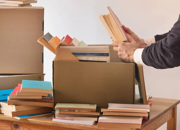 Two man hands are packing books in a cardboard box ,table with stacks of books ,two boxes on the left background, daylight ,white background