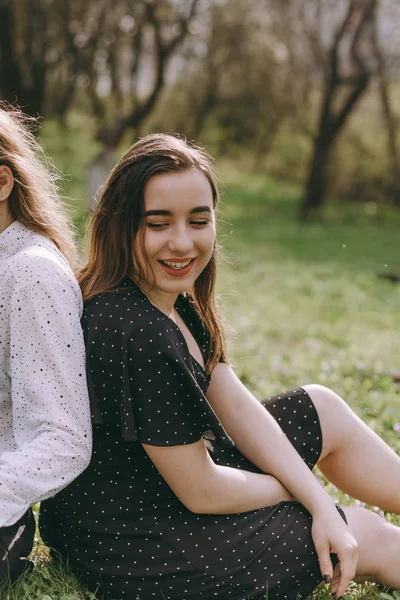 Girl sitting smiling and leaning on her sister in the green spring garden, hugging in the field in the sunshine. Having fun together, positive emotions, bright colors. Happy girlfriends at sunset — Stock Photo, Image