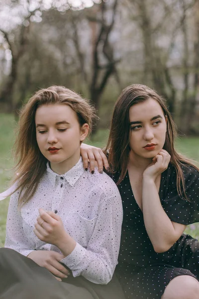 Portrait of two beautiful young sisters sitting in the green spring park, hugging in the field. Having fun together, positive emotions, bright colors. Copy space. Happy girlfriends at sunset — Stock Photo, Image