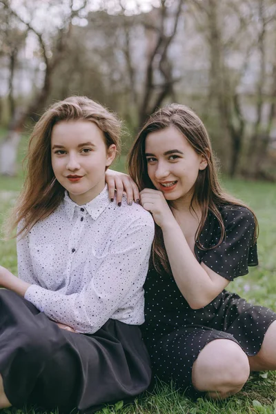 Porträt von zwei schönen jungen Schwestern, die im grünen Frühlingspark sitzen und sich auf dem Feld umarmen. Gemeinsam Spaß haben, positive Emotionen, bunte Farben. Kopierraum. glückliche Freundinnen bei Sonnenuntergang — Stockfoto