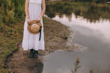 Gün batımında göl kenarında duran güzel boho kızı. Beyaz bohem elbiseli, rüzgarlı saçlı, yeşil yaprakların arasında, suyun yanında dinlenen hasır şapkalı çekici genç bir kadın. yaz tatili