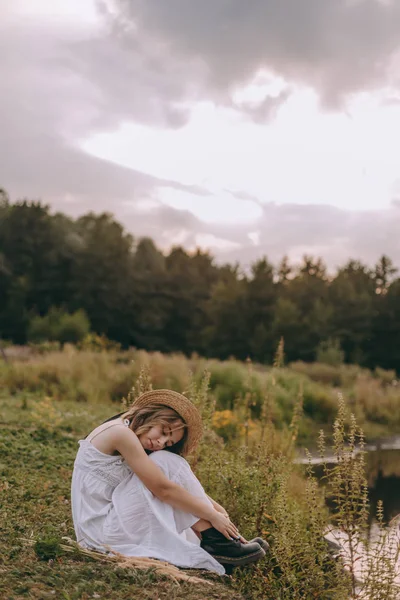 güzel boho kız göl yakınında gün batımı ışıkta oturan. yeşil yaprakları arasında su yakınında rahatlatıcı rüzgarlı saçları ile beyaz bohem elbiseli çekici genç kadın. yaz tatili
