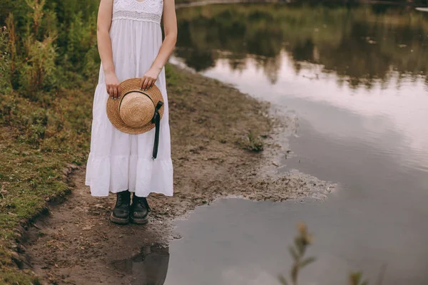 Красива дівчина-бухо, що стоїть на заході сонця біля озера. приваблива молода жінка в білій богемній сукні з вітряним волоссям, що тримає солом'яний капелюх, що розслабляється біля води серед зеленого листя. літні канікули — стокове фото