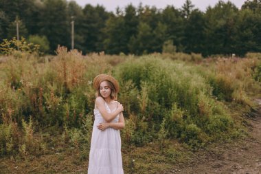 Gün batımında göl kenarında duran güzel boho kızı. Beyaz bohem elbiseli, rüzgarlı saçlı, yeşil yaprakların arasında, suyun yanında dinlenen hasır şapkalı çekici genç bir kadın. yaz tatili